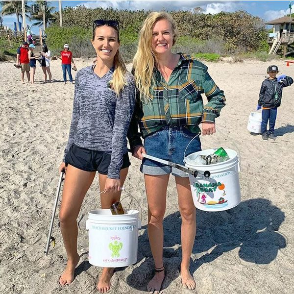 Two smiling volunteers stand barefoot on a sunny beach, holding trash grabbers and white buckets partially filled with collected debris. The buckets feature the Beach Bucket Foundation logo, with other cleanup participants visible in the background.
