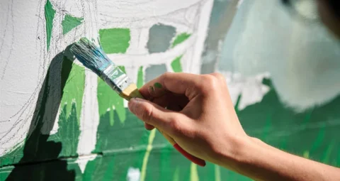 A close-up of a hand painting a green outdoor mural.