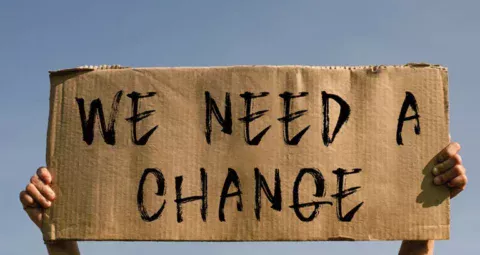 A cardboard protest sign reading 'WE NEED A CHANGE'.