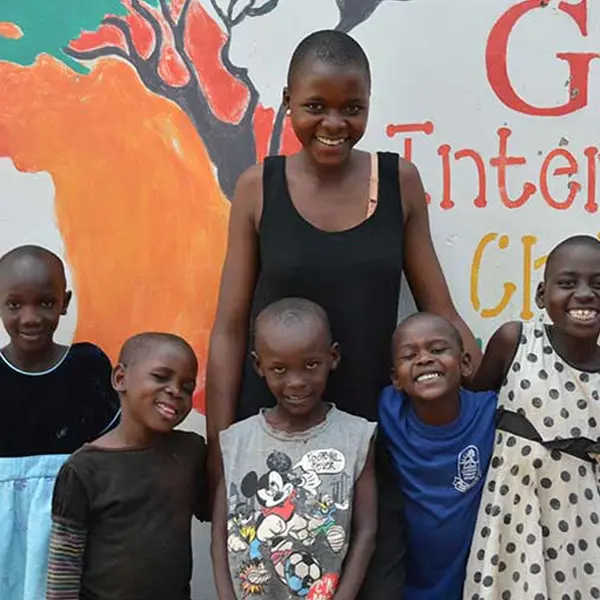 A smiling young woman and five happy children stand together in front of a colorful wall mural that features a map of Africa and partial lettering for the foundation.