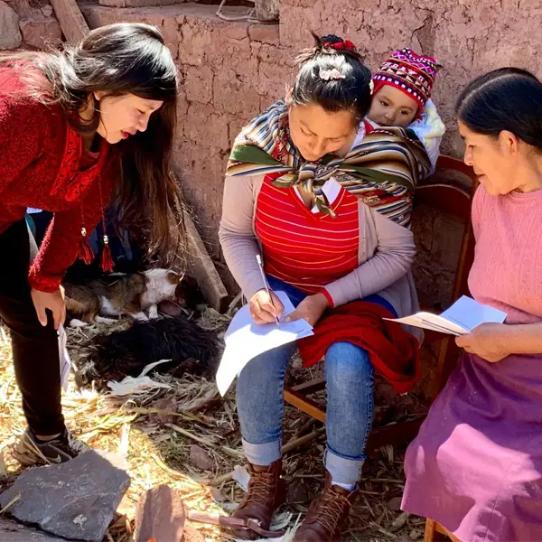 A woman carrying a baby on her back in a colorful woven cloth sits outdoors writing on paperwork, closely assisted by a standing woman in red, while a third woman holding papers sits nearby
