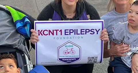 Two smiling women and two children, one seated in a specialized stroller, posing together at an outdoor event. The woman in the center is kneeling and holding a sign that reads 'KCNT1 EPILEPSY FOUNDATION, RIDE FOR RESEARCH, TEAM KCNT1'.