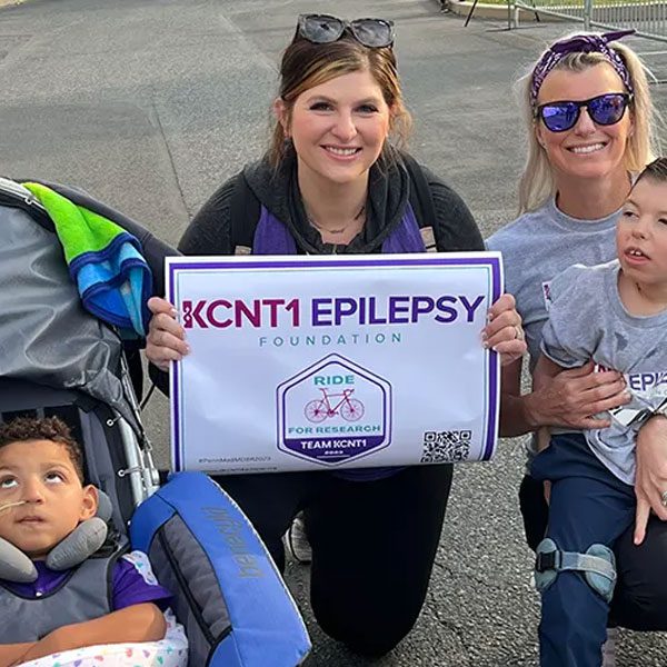 Two smiling women and two children, one seated in a specialized stroller, posing together at an outdoor event. The woman in the center is kneeling and holding a sign that reads 'KCNT1 EPILEPSY FOUNDATION, RIDE FOR RESEARCH, TEAM KCNT1'.