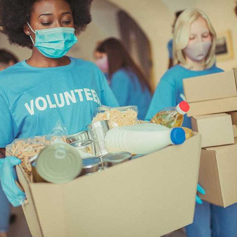 A volunteer wearing a blue 'VOLUNTEER' shirt, face mask, and gloves carries a large cardboard box filled with donated food items like canned goods and a milk jug, while another volunteer carries stacked boxes in the background.