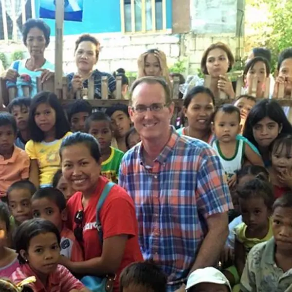 A smiling man and woman stand surrounded by a large, joyful group of children and community members gathered outdoors in a local neighborhood.