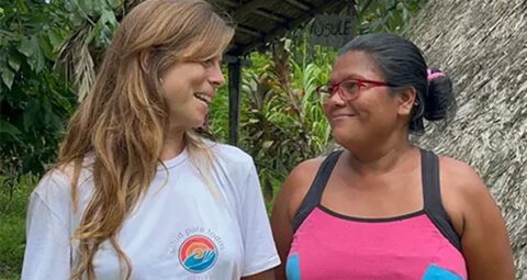 A smiling woman wearing a white 'Salud Para Todos Caribe' t-shirt happily converses with another smiling woman outdoors in a tropical setting near a thatched-roof structure.