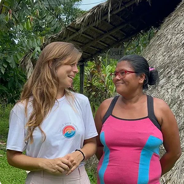 A smiling woman wearing a white 'Salud Para Todos Caribe' t-shirt happily converses with another smiling woman outdoors in a tropical setting near a thatched-roof structure.