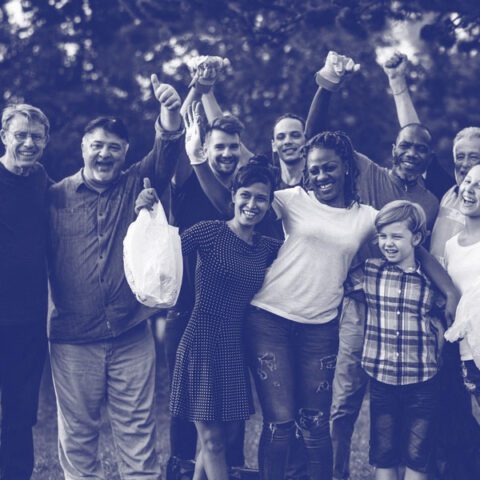 A large, diverse group of volunteers of all ages standing outdoors in a park, smiling and cheering with their hands raised. Several people in the group are wearing work gloves and holding white garbage bags. The photo has a stylized blue monochromatic tint.