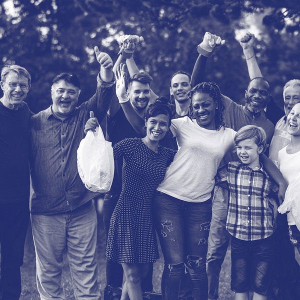 A large, diverse group of volunteers of all ages standing outdoors in a park, smiling and cheering with their hands raised. Several people in the group are wearing work gloves and holding white garbage bags. The photo has a stylized blue monochromatic tint.